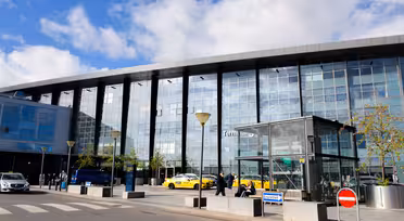 Udvendig udsigt af en moderne lufthavnsterminal med en glasfacade. Flere gule taxaer og et par personer står foran indgangen. Himlen er delvist overskyet, og der er træer og gadeskilte i nærheden af terminalen.