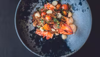 En mørk skål fyldt med en farverig ret med levende rød hummer, cherrytomater, hvide bønner og hakkede krydderurter, alt sammen blandet i en krydret sauce, er sat på en mørk baggrund.