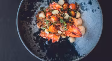 En mørk skål fyldt med en farverig ret med levende rød hummer, cherrytomater, hvide bønner og hakkede krydderurter, alt sammen blandet i en krydret sauce, er sat på en mørk baggrund.