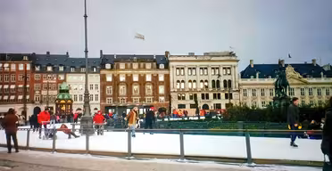Folk, der skøjter på en udendørs skøjtebane på et bytorv, omgivet af historiske bygninger i europæisk stil. En statue og lygtepæl er synlige, og nogle mennesker ser på fra sidelinjen. Det ser ud til at være dagtimerne.