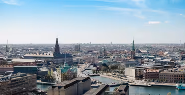 Luftfoto af en europæisk by med historiske bygninger, en snoet flod og flere strukturer med spir. Den blå himmel over er let skyet, og bylandskabet strækker sig til horisonten.