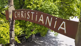 En buegang i træ med "Christiania" skrevet med store hvide bogstaver. Buen har udskårne ansigter på siderne. Frodige grønne træer omgiver området, og cykler er parkeret nedenfor. Scenen er solbelyst, hvilket skaber en levende og livlig atmosfære.