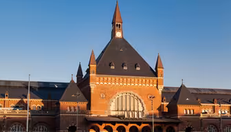 En stor murstensbygning med et højt, centralt klokketårn og trekantet tag, med buede vinduer og detaljeret murværk. Facaden er symmetrisk med mindre spir på hver side, under en klar blå himmel.