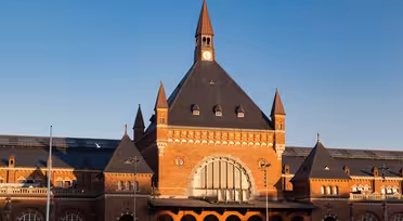 En stor murstensbygning med et højt, centralt klokketårn og trekantet tag, med buede vinduer og detaljeret murværk. Facaden er symmetrisk med mindre spir på hver side, under en klar blå himmel.