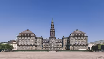 Storslået historisk bygning under en klar blå himmel med udsmykket arkitektur og et centralt tårn. Forgrunden omfatter en rummelig, tom plads med pænt trimmede træer langs siderne.