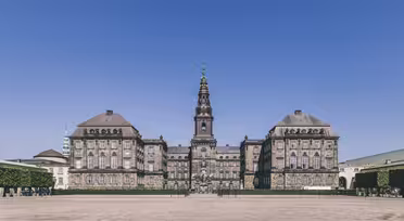 Storslået historisk bygning under en klar blå himmel med udsmykket arkitektur og et centralt tårn. Forgrunden omfatter en rummelig, tom plads med pænt trimmede træer langs siderne.