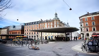 En bygadescene med et moderne, cirkulært baldakinskjul i centrum, omgivet af historiske bygninger i europæisk stil. Adskillige cykler er parkeret, og enkelte fodgængere og cyklister er synlige. En faldet cykel ligger på fortovet.