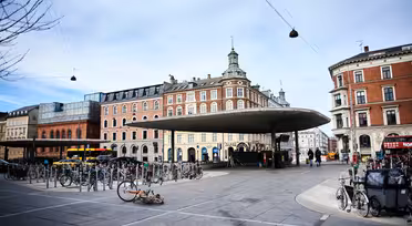 En bygadescene med et moderne, cirkulært baldakinskjul i centrum, omgivet af historiske bygninger i europæisk stil. Adskillige cykler er parkeret, og enkelte fodgængere og cyklister er synlige. En faldet cykel ligger på fortovet.