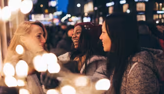 Tre kvinder smiler og taler i en festlig, oplyst gade om natten. De er iført vintertøj, inklusive frakker og en rød hue. Sløret lys skaber en varm forgrund, der forstærker den muntre, hyggelige atmosfære.