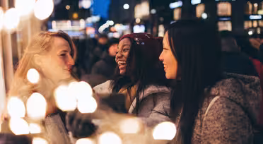 Tre kvinder smiler og taler i en festlig, oplyst gade om natten. De er iført vintertøj, inklusive frakker og en rød hue. Sløret lys skaber en varm forgrund, der forstærker den muntre, hyggelige atmosfære.