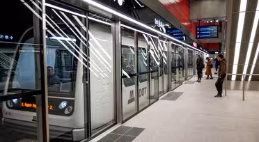 Et moderne metrotog stoppede ved en station. Passagerer er synlige inde i toget, mens et par personer venter på perronen. Stationen er lys med overheadbelysning og digitale skilte, der viser information.