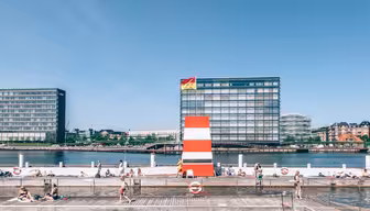 Folk svømmer og slapper af ved en pool ved vandet med klart vand og træterrasser. I baggrunden står moderne bygninger og en stor rød-hvid-stribet dykkerplatform mod en blå himmel.