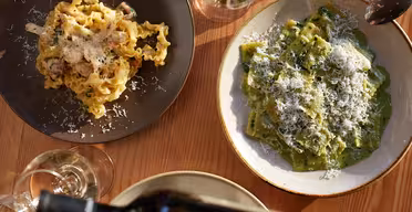 To plader pasta på et træbord: den ene med mafaldinepasta og den anden med grøn ravioli, begge toppet med revet ost. En person hælder vin i et glas i forgrunden. Briller og en ske er i nærheden.