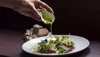 En hånd hælder grøn sauce fra en østersskal på en hvid tallerken med bladgrønt, urter og skiver speget kød. Et par østersskaller er i baggrunden.