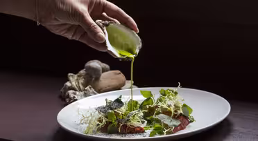 En hånd hælder grøn sauce fra en østersskal på en hvid tallerken med bladgrønt, urter og skiver speget kød. Et par østersskaller er i baggrunden.
