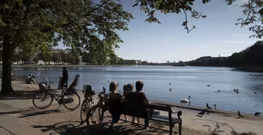 Folk sidder på en bænk ved en fredfyldt sø i træskygge. Cykler er parkeret i nærheden. Ænder og svaner flyder på vandet. Scenen omfatter fjerne bygninger og en klar blå himmel.