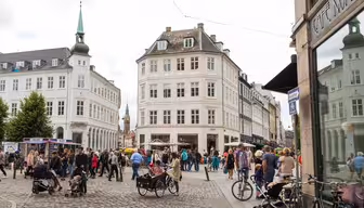 Et travlt europæisk gadebillede med folk, der går, cykler og sidder uden for caféer. Området byder på brostensbelagte gader, historiske bygninger med høje vinduer og spir i baggrunden under en overskyet himmel.