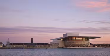 Billedet viser en udsigt over vandet af et moderne operahus med et unikt kantet tag, sat mod en lyserød og lilla skumringshimmel. Vandet i forgrunden reflekterer lyset fra himlen, hvilket skaber en fredelig scene.
