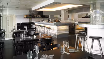 Moderne restaurantinteriør med sorte borde og stole, elegant sat med glas og servietter. En veloplyst bar med taburetter er i baggrunden, og loftet har et industrielt design. Stemningen er slank og moderne.