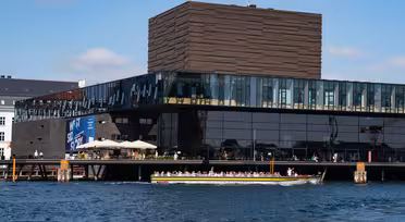 En stor moderne bygning med reflekterende glasvinduer og en struktureret brun facade ligger ved en vandmasse. En kanalbåd med passagerer passerer foran bygningen under en blå himmel.