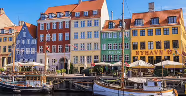 Farverige historiske bygninger ligger langs havnefronten ved Nyhavn i København, med sejlbåde fortøjet langs kanalen. De lyse facader af røde, gule og grønne bygninger spejler sig i vandet under en klar blå himmel.