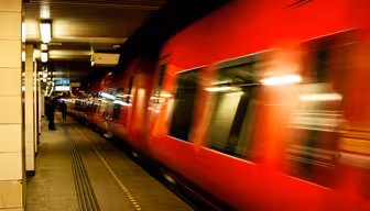 Et knaldrødt tog suser gennem en svagt oplyst underjordisk station og skaber en bevægelsessløring. Platformen er tom bortset fra nogle få fjerne figurer. Overheadlys og prikkede platformsmarkeringer er synlige.