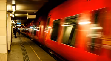 Et knaldrødt tog suser gennem en svagt oplyst underjordisk station og skaber en bevægelsessløring. Platformen er tom bortset fra nogle få fjerne figurer. Overheadlys og prikkede platformsmarkeringer er synlige.