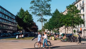 En solrig byscene viser to mennesker, der går med en cykel på en brostensbelagt sti. Træer og bygninger ligger langs gaden. Folk sidder og samles på græsarealet og nyder det behagelige vejr. Biler er parkeret langs vejen.