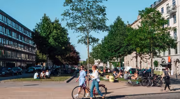 En solrig byscene viser to mennesker, der går med en cykel på en brostensbelagt sti. Træer og bygninger ligger langs gaden. Folk sidder og samles på græsarealet og nyder det behagelige vejr. Biler er parkeret langs vejen.
