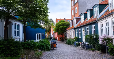 En charmerende brostensbelagt gade med maleriske huse med farverigt ydre og skrå tage. Cykler er parkeret langs siden, og frodige grønne områder, herunder træer og buske, tilføjer et naturligt præg til den fredfyldte, overskyede scene.