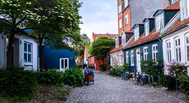 En charmerende brostensbelagt gade med maleriske huse med farverigt ydre og skrå tage. Cykler er parkeret langs siden, og frodige grønne områder, herunder træer og buske, tilføjer et naturligt præg til den fredfyldte, overskyede scene.