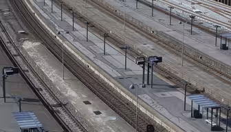 En tom jernbanestation med flere parallelle spor og perroner. Adskillige skilte og shelters med bænke er synlige langs perronerne. Scenen er godt oplyst, hvilket tyder på dagtimerne, uden tog eller mennesker i syne.