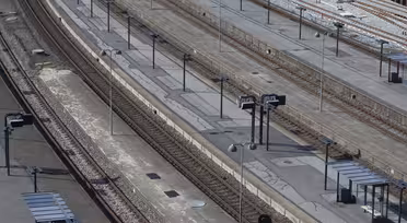 En tom jernbanestation med flere parallelle spor og perroner. Adskillige skilte og shelters med bænke er synlige langs perronerne. Scenen er godt oplyst, hvilket tyder på dagtimerne, uden tog eller mennesker i syne.