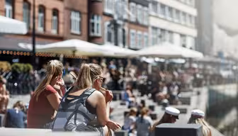 Folk, der sidder udendørs ved en havnefront på en solskinsdag. Nogle er iført sommertøj, og enkelte er iklædt sømandshatte, muligvis dimittender. Baggrunden byder på bygninger med caféer og paraplyer med en livlig, afslappet atmosfære.