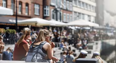 Folk, der sidder udendørs ved en havnefront på en solskinsdag. Nogle er iført sommertøj, og enkelte er iklædt sømandshatte, muligvis dimittender. Baggrunden byder på bygninger med caféer og paraplyer med en livlig, afslappet atmosfære.