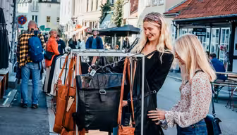To kvinder gennemser lædertasker på et gademarkedsstativ. De er smilende og engagerede, mens andre går og shopper i baggrunden. Det er en solskinsdag, og markedet ligger i en malerisk, travl by med charmerende bygninger.
