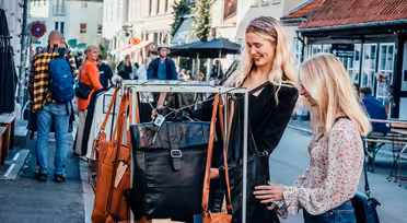 To kvinder gennemser lædertasker på et gademarkedsstativ. De er smilende og engagerede, mens andre går og shopper i baggrunden. Det er en solskinsdag, og markedet ligger i en malerisk, travl by med charmerende bygninger.