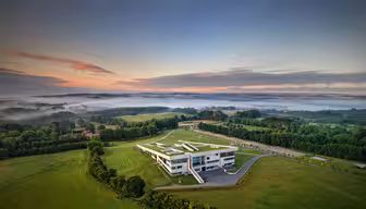 Luftfoto af en moderne bygning med et geometrisk design beliggende i et frodigt grønt landskab. Tåge dækker dele af landskabet. Himlen viser en gradient fra orange til blå, når solen går ned eller står op.