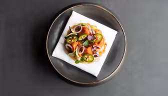 En gourmetret med et rundt stykke toast toppet med skåret jalapeños, løg, mikrogrønt og spiselige blomster. Den placeres på en hvid serviet inde i en sort skål mod en mørkegrå baggrund.
