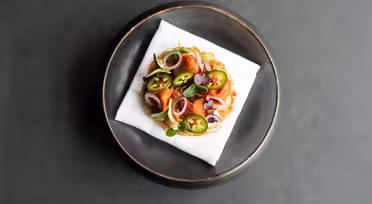 En gourmetret med et rundt stykke toast toppet med skåret jalapeños, løg, mikrogrønt og spiselige blomster. Den placeres på en hvid serviet inde i en sort skål mod en mørkegrå baggrund.