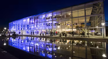 Moderne glasbygning oplyst med levende lilla og blå lys om natten, der reflekterer på en rolig vandoverflade i forgrunden. Folk samles omkring den veloplyste indgang og skaber en livlig atmosfære.