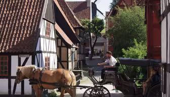 En hestevogn bevæger sig gennem en brostensbelagt gade i en hyggelig landsby med bindingsværkshuse og en vindmølle i baggrunden. Scenen er solrig og fredelig.