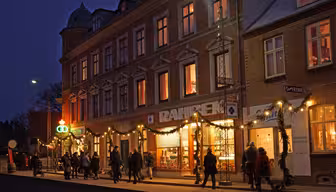 En gruppe mennesker går langs en gade foran en varmt oplyst bygning prydet med festlige lys og guirlander om aftenen. Atmosfæren er livlig og indbydende, med butikker og dekorative vinduer synlige.