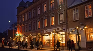 En gruppe mennesker går langs en gade foran en varmt oplyst bygning prydet med festlige lys og guirlander om aftenen. Atmosfæren er livlig og indbydende, med butikker og dekorative vinduer synlige.