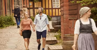 Et par holder i hånden, mens de går langs en brostensbelagt gade med træbygninger. Manden har en lys skjorte og shorts på, og kvinden har en pink top og sort nederdel på. En kvinde i traditionel påklædning går foran. Frodigt grønt omgiver området.