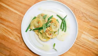 En hvid tallerken med ravioli overhældt med en let sauce, garneret med grønne bønner og pinjekerner. Et par basilikumblade er spredt ovenpå. Pladen er sat på en træoverflade.