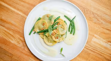 En hvid tallerken med ravioli overhældt med en let sauce, garneret med grønne bønner og pinjekerner. Et par basilikumblade er spredt ovenpå. Pladen er sat på en træoverflade.