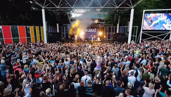 En stor folkemængde ser et band optræde på en udendørs scene. Farverige lodrette striber dekorerer sceneområdet. Røgeffekter og skarpt lys forbedrer ydeevnen, når folk holder hænderne op i begejstring.