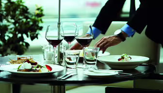 En person i jakkesæt dækker et bord med tallerkener med gourmetmad, vinglas fyldt med rødvin og vandglas. Bordet er elegant arrangeret i moderne restaurant-miljø med en plante i baggrunden.