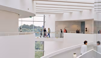 Moderne arkitektonisk interiør med buet trappe og store glasvinduer. Folk går på det øverste niveau med udsigt over grønne områder udenfor. Rummet er lyst med et minimalistisk hvidt design.